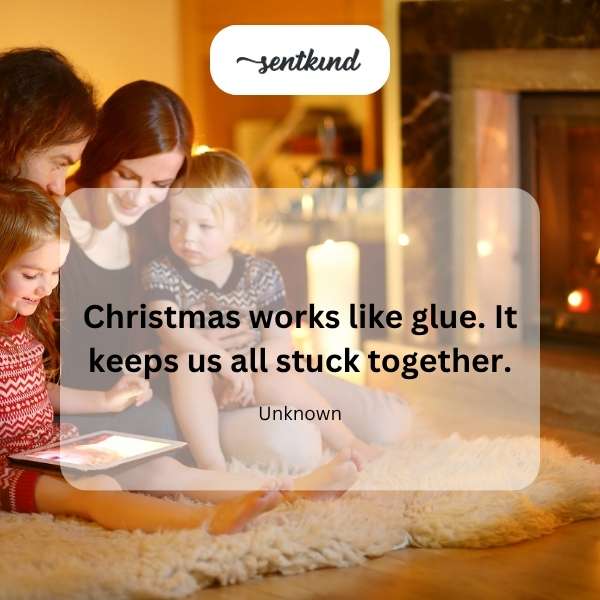 A diverse group of people are gathered closely together, some holding gifts, with a softly glowing Christmas tree in the background.