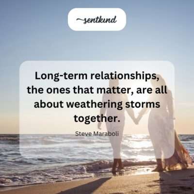 A couple stands together on a rocky shore, weathered but strong, looking out at a stormy sea with hands clasped.