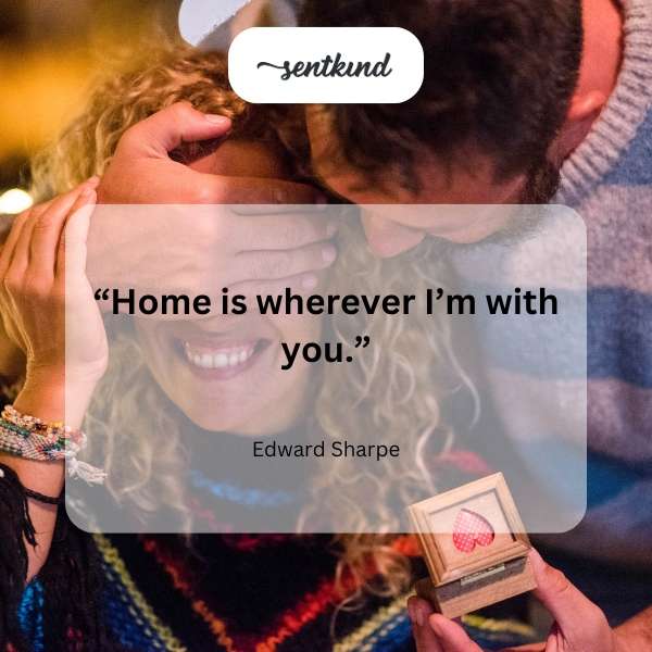 A couple embraces warmly in front of a cozy, warmly lit house.